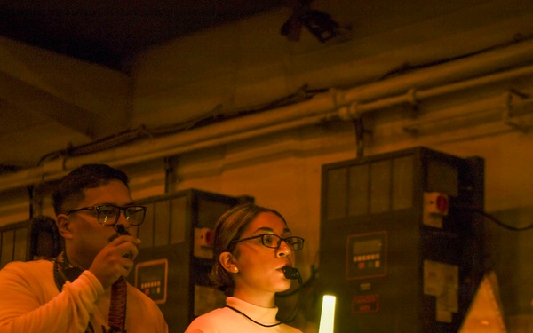 Two individuals are standing in a dimly lit industrial or mechanical space, illuminated primarily by warm orange emergency lighting and a glowing chemical light stick held by the woman on the right. Both are wearing matching sweatshirts with partial text visible ('PO' and 'HGR'). They appear to be conducting or participating in some kind of drill or operational exercise.