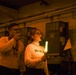 Two individuals are standing in a dimly lit industrial or mechanical space, illuminated primarily by warm orange emergency lighting and a glowing chemical light stick held by the woman on the right. Both are wearing matching sweatshirts with partial text visible ('PO' and 'HGR'). They appear to be conducting or participating in some kind of drill or operational exercise.