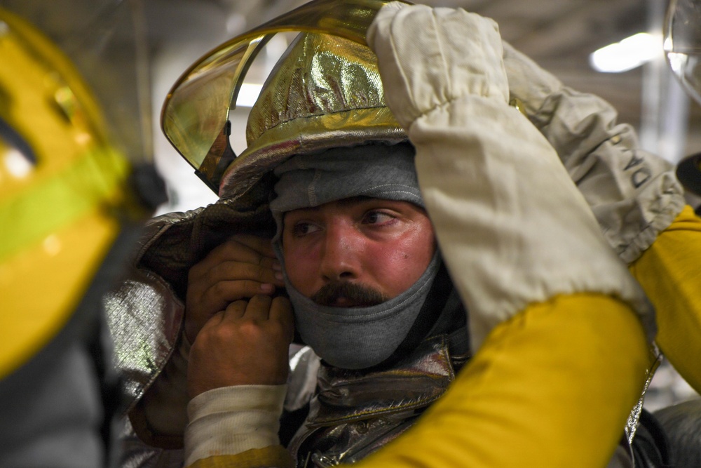 A firefighter is being assisted in donning or adjusting proximity/aluminized firefighting gear, including a gold reflective helmet with a visor. The subject appears to be in a training or preparation environment, with visible signs of heat stress on his face. Another person in yellow protective gear is helping secure the equipment.