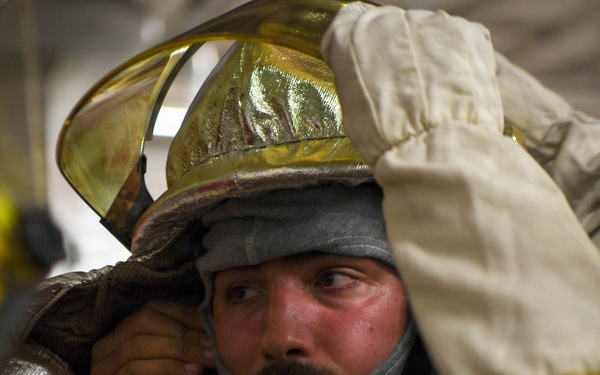 A firefighter is being assisted in donning or adjusting proximity/aluminized firefighting gear, including a gold reflective helmet with a visor. The subject appears to be in a training or preparation environment, with visible signs of heat stress on his face. Another person in yellow protective gear is helping secure the equipment.
