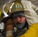 A firefighter is being assisted in donning or adjusting proximity/aluminized firefighting gear, including a gold reflective helmet with a visor. The subject appears to be in a training or preparation environment, with visible signs of heat stress on his face. Another person in yellow protective gear is helping secure the equipment.