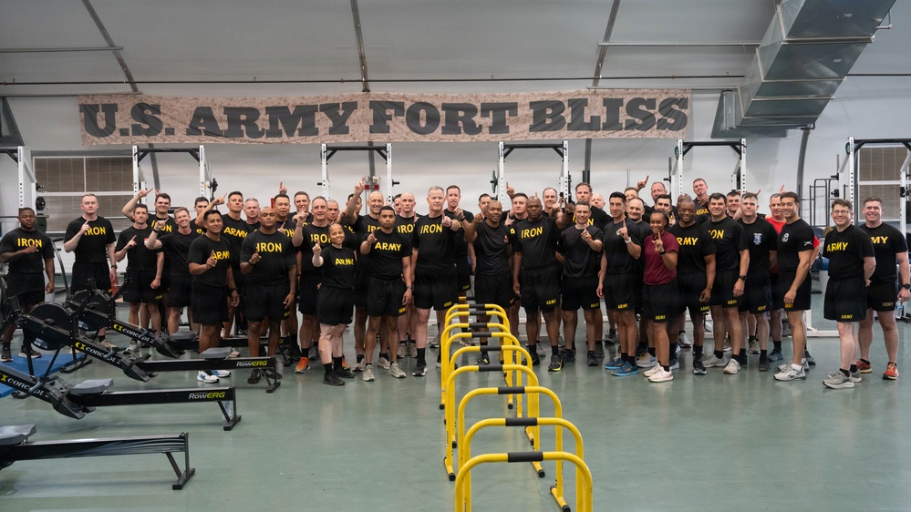 A large group of U.S. Army personnel pose for a group photo inside a fitness facility at Fort Bliss. The individuals are wearing black Army physical training uniforms with 'IRON' and 'ARMY' text, and many are giving a thumbs-up gesture. The gym is equipped with rowing machines and yellow parallel bars/hurdles in the foreground.