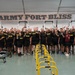 A large group of U.S. Army personnel pose for a group photo inside a fitness facility at Fort Bliss. The individuals are wearing black Army physical training uniforms with 'IRON' and 'ARMY' text, and many are giving a thumbs-up gesture. The gym is equipped with rowing machines and yellow parallel bars/hurdles in the foreground.