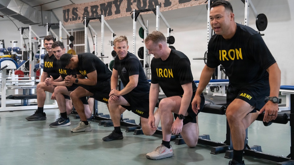 Un grupo de soldados del ejército estadounidense están reunidos en un gimnasio militar, vistiendo camisetas y pantalones cortos negros de entrenamiento físico 'IRON ARMY' a juego. Parecen estar arrodillados o arremetiendo, posiblemente recibiendo instrucción o participando en una actividad física grupal. El escenario es claramente un gimnasio de instalación del ejército de EE. UU., como lo indica el letrero que dice 