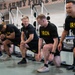 A group of U.S. Army soldiers are gathered in a military gym facility, wearing matching black 'IRON ARMY' physical training shirts and shorts. They appear to be in a kneeling or lunging position, possibly receiving instruction or participating in a group fitness activity. The setting is clearly a U.S. Army installation gym, as indicated by the 'U.S. ARMY FORT' signage visible on the wall.