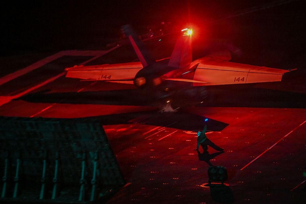 A night operations scene on what appears to be an aircraft carrier flight deck, showing a military jet aircraft (marked '144' on both wings) under dramatic red lighting. A flight deck crew member is visible in the foreground, illuminated by a blue light indicator. The scene captures the high-intensity environment of nocturnal carrier flight operations.