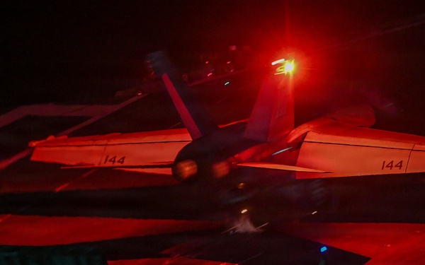 A night operations scene on what appears to be an aircraft carrier flight deck, showing a military jet aircraft (marked '144' on both wings) under dramatic red lighting. A flight deck crew member is visible in the foreground, illuminated by a blue light indicator. The scene captures the high-intensity environment of nocturnal carrier flight operations.