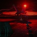 A night operations scene on what appears to be an aircraft carrier flight deck, showing a military jet aircraft (marked '144' on both wings) under dramatic red lighting. A flight deck crew member is visible in the foreground, illuminated by a blue light indicator. The scene captures the high-intensity environment of nocturnal carrier flight operations.