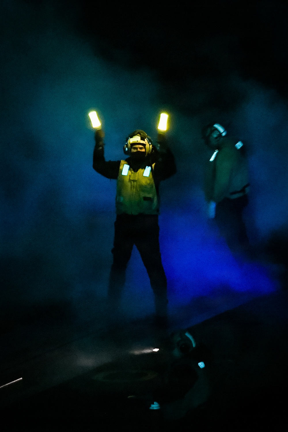 A person wearing a yellow/green safety vest and helmet with hearing protection stands in a dark, smoke-filled environment holding two illuminated light wands raised above their head. A second individual is partially visible on the right side of the frame. The scene is consistent with night flight deck operations aboard a naval vessel, where aircraft marshallers use lighted wands to guide aircraft.