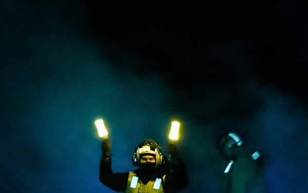 A person wearing a yellow/green safety vest and helmet with hearing protection stands in a dark, smoke-filled environment holding two illuminated light wands raised above their head. A second individual is partially visible on the right side of the frame. The scene is consistent with night flight deck operations aboard a naval vessel, where aircraft marshallers use lighted wands to guide aircraft.