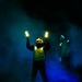 A person wearing a yellow/green safety vest and helmet with hearing protection stands in a dark, smoke-filled environment holding two illuminated light wands raised above their head. A second individual is partially visible on the right side of the frame. The scene is consistent with night flight deck operations aboard a naval vessel, where aircraft marshallers use lighted wands to guide aircraft.