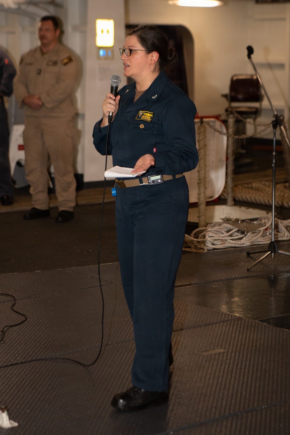 A female military officer in dark navy blue coveralls is speaking into a handheld microphone while holding papers, appearing to address an audience. She is standing on what appears to be the interior deck of a naval vessel, identifiable by the non-slip flooring, mooring ropes, and watertight door in the background. A second person in tan/khaki military uniform is visible standing in the background.