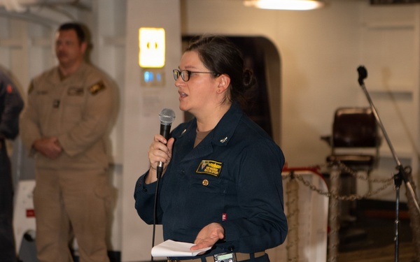 A female military officer in dark navy blue coveralls is speaking into a handheld microphone while holding papers, appearing to address an audience. She is standing on what appears to be the interior deck of a naval vessel, identifiable by the non-slip flooring, mooring ropes, and watertight door in the background. A second person in tan/khaki military uniform is visible standing in the background.