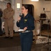A female military officer in dark navy blue coveralls is speaking into a handheld microphone while holding papers, appearing to address an audience. She is standing on what appears to be the interior deck of a naval vessel, identifiable by the non-slip flooring, mooring ropes, and watertight door in the background. A second person in tan/khaki military uniform is visible standing in the background.