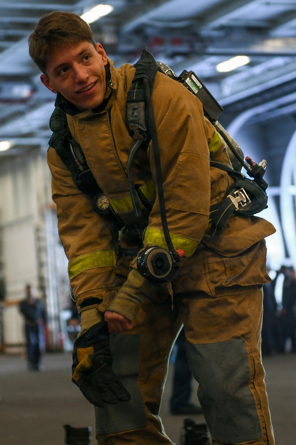 A young male firefighter or damage control personnel is wearing full turnout gear including a tan/gold bunker suit with reflective yellow-green trim and an SCBA (Self-Contained Breathing Apparatus) on his back. He is smiling at the camera while appearing to don or adjust his equipment. The setting appears to be the hangar bay of a naval vessel, indicated by the large curved interior structure and overhead lighting.