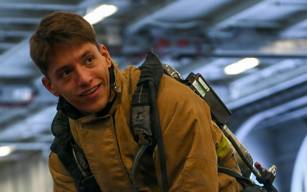 A young male firefighter or damage control personnel is wearing full turnout gear including a tan/gold bunker suit with reflective yellow-green trim and an SCBA (Self-Contained Breathing Apparatus) on his back. He is smiling at the camera while appearing to don or adjust his equipment. The setting appears to be the hangar bay of a naval vessel, indicated by the large curved interior structure and overhead lighting.