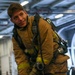 A young male firefighter or damage control personnel is wearing full turnout gear including a tan/gold bunker suit with reflective yellow-green trim and an SCBA (Self-Contained Breathing Apparatus) on his back. He is smiling at the camera while appearing to don or adjust his equipment. The setting appears to be the hangar bay of a naval vessel, indicated by the large curved interior structure and overhead lighting.