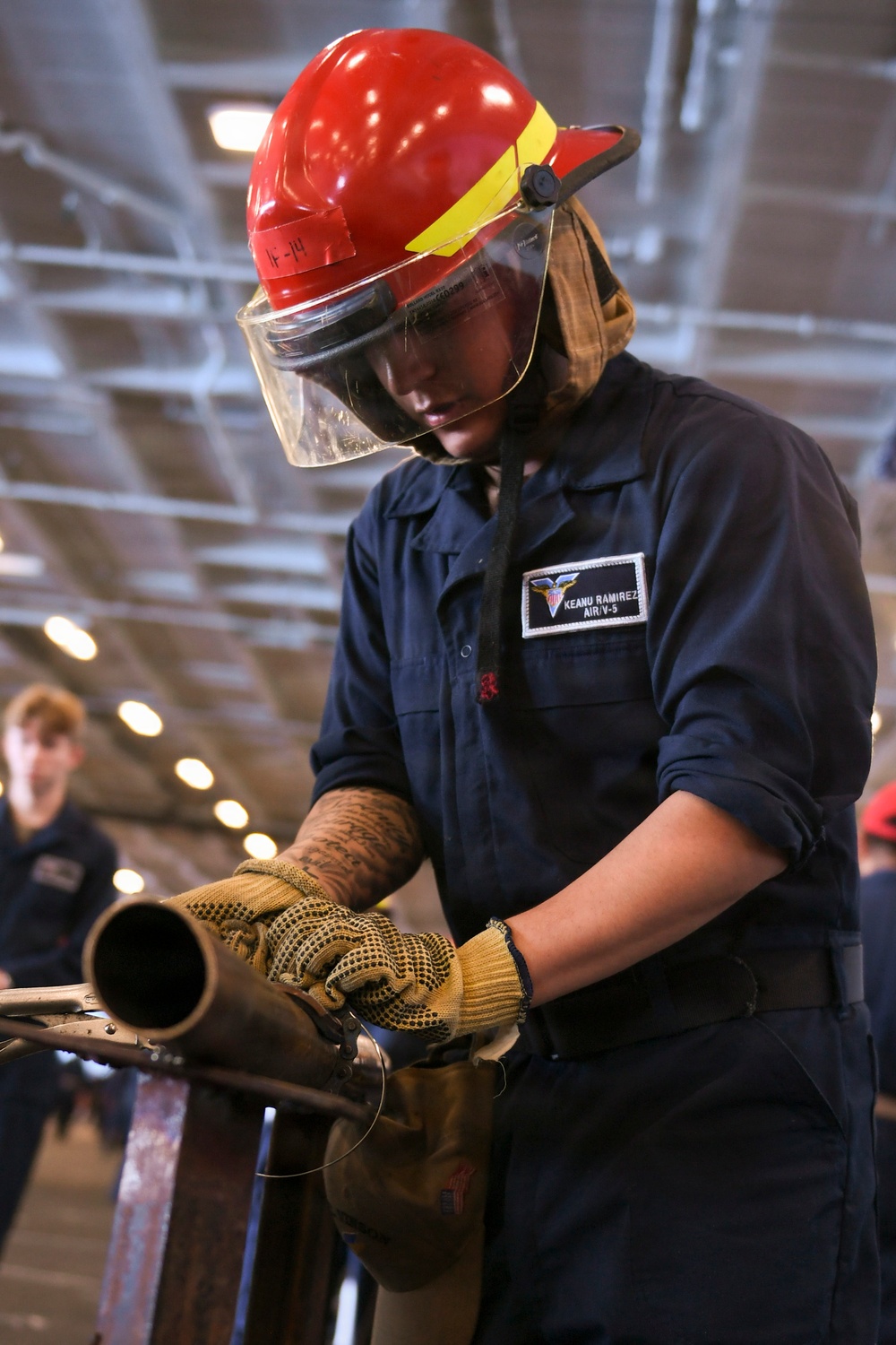 A U.S. Navy sailor identified as Keanu Ramirez from AIR/V-5 department is handling a fire hose nozzle during what appears to be a damage control or firefighting drill aboard a naval vessel. The sailor is wearing a red firefighting helmet with a clear face shield, navy blue coveralls, and yellow Kevlar work gloves. The setting appears to be inside a ship's hangar bay based on the overhead lighting and structural elements visible in the background.