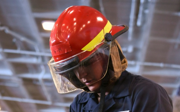 A U.S. Navy sailor identified as Keanu Ramirez from AIR/V-5 department is handling a fire hose nozzle during what appears to be a damage control or firefighting drill aboard a naval vessel. The sailor is wearing a red firefighting helmet with a clear face shield, navy blue coveralls, and yellow Kevlar work gloves. The setting appears to be inside a ship's hangar bay based on the overhead lighting and structural elements visible in the background.