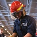 A U.S. Navy sailor identified as Keanu Ramirez from AIR/V-5 department is handling a fire hose nozzle during what appears to be a damage control or firefighting drill aboard a naval vessel. The sailor is wearing a red firefighting helmet with a clear face shield, navy blue coveralls, and yellow Kevlar work gloves. The setting appears to be inside a ship's hangar bay based on the overhead lighting and structural elements visible in the background.