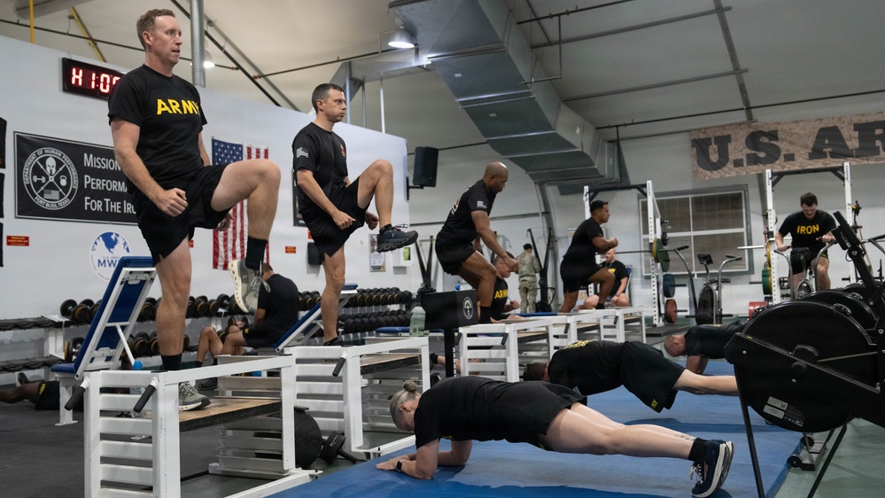 A group of U.S. Army soldiers are engaged in a structured physical training session inside a military fitness facility. Multiple exercises are being performed simultaneously, including high-knee step-ups on white plyometric boxes, push-ups on blue mats, and stationary bike cycling. The setting appears to be an official Army gym, indicated by 'ARMY' text on uniforms and 'U.S. AF' signage on the wall.