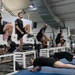 A group of U.S. Army soldiers are engaged in a structured physical training session inside a military fitness facility. Multiple exercises are being performed simultaneously, including high-knee step-ups on white plyometric boxes, push-ups on blue mats, and stationary bike cycling. The setting appears to be an official Army gym, indicated by 'ARMY' text on uniforms and 'U.S. AF' signage on the wall.