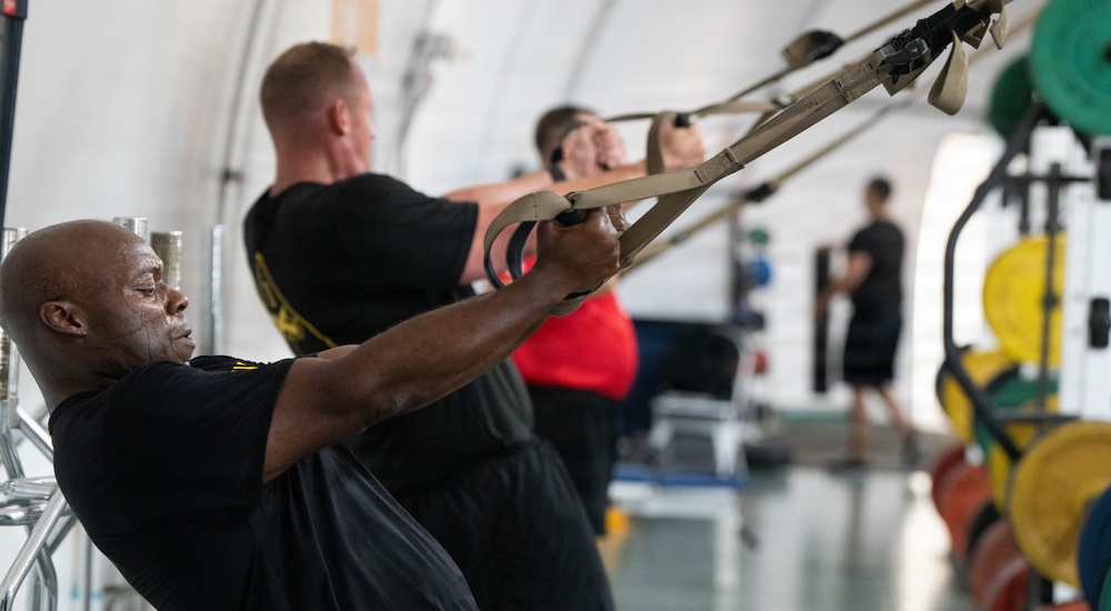 A group of individuals are performing TRX suspension training exercises in what appears to be a military or institutional gym facility. Multiple participants are leaning back while gripping suspension straps, engaging their core and upper body muscles. The foreground subject is a Black male in a black t-shirt showing visible exertion, with additional participants lined up behind him in similar positions.