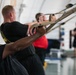 A group of individuals are performing TRX suspension training exercises in what appears to be a military or institutional gym facility. Multiple participants are leaning back while gripping suspension straps, engaging their core and upper body muscles. The foreground subject is a Black male in a black t-shirt showing visible exertion, with additional participants lined up behind him in similar positions.