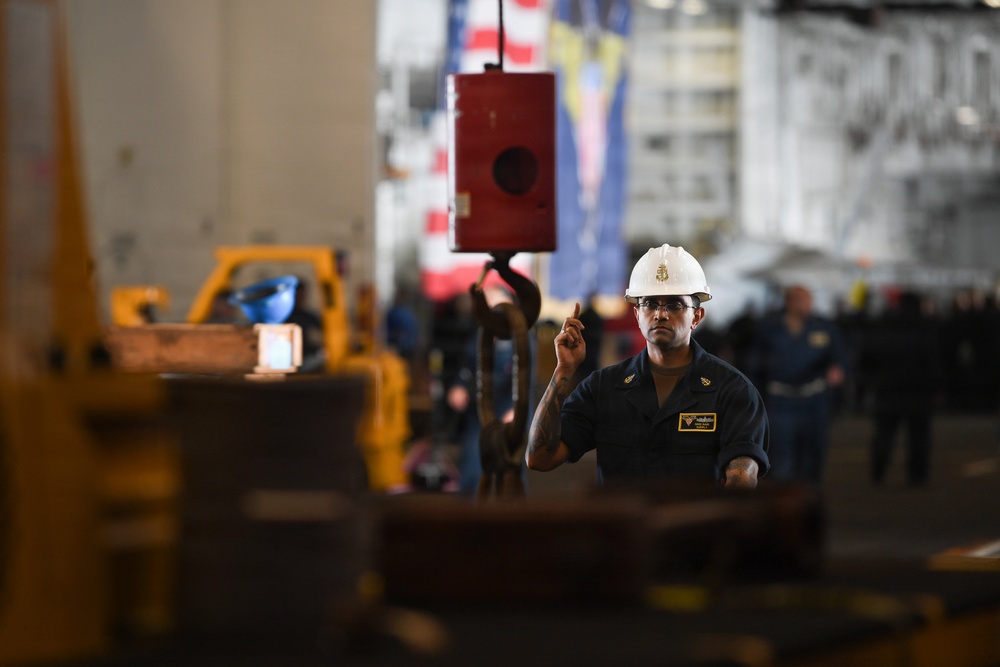 A worker wearing a white hard hat and dark navy coveralls is directing or signaling with one finger raised, likely guiding a crane or hoist operation in an industrial or military hangar bay setting. A red crane hook block is suspended in the foreground, and an American flag along with another flag are visible in the background. The environment appears to be a large enclosed facility, possibly a naval vessel hangar deck.