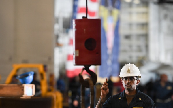 A worker wearing a white hard hat and dark navy coveralls is directing or signaling with one finger raised, likely guiding a crane or hoist operation in an industrial or military hangar bay setting. A red crane hook block is suspended in the foreground, and an American flag along with another flag are visible in the background. The environment appears to be a large enclosed facility, possibly a naval vessel hangar deck.
