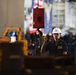 A worker wearing a white hard hat and dark navy coveralls is directing or signaling with one finger raised, likely guiding a crane or hoist operation in an industrial or military hangar bay setting. A red crane hook block is suspended in the foreground, and an American flag along with another flag are visible in the background. The environment appears to be a large enclosed facility, possibly a naval vessel hangar deck.