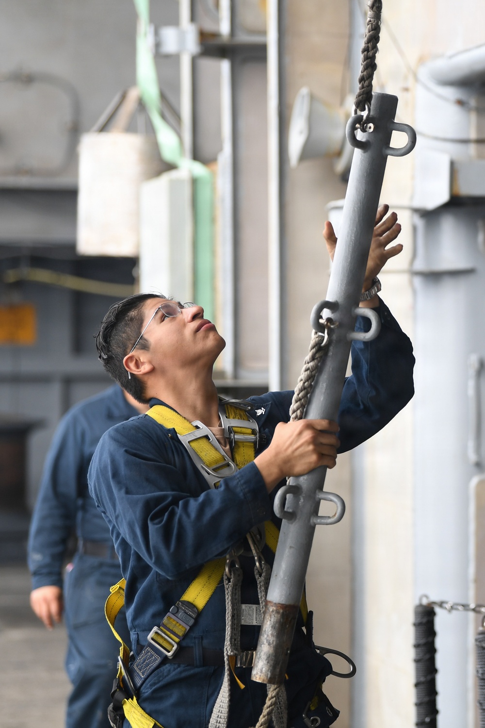 A person wearing a navy blue coverall uniform and yellow safety harness is handling a metal pole or stanchion with a rope attached, looking upward in what appears to be a naval vessel or industrial facility. The individual is wearing safety glasses and appears to be performing a rigging or equipment installation task. A second person in similar attire is visible in the background.