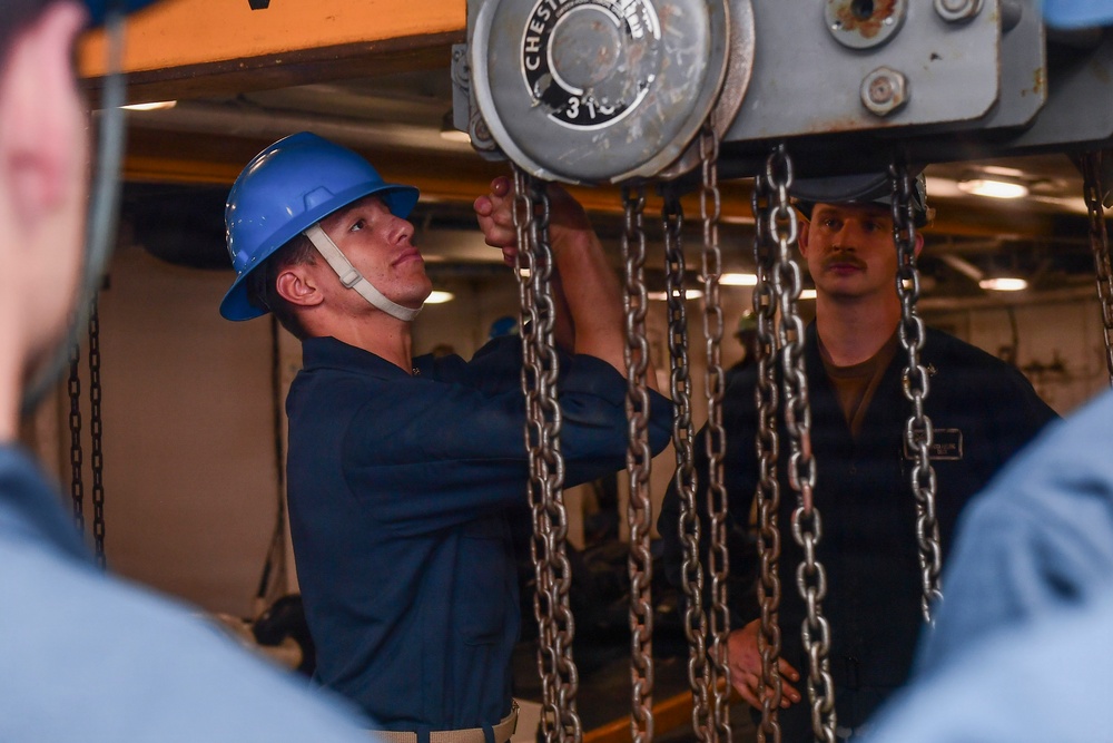 Two personnel in dark navy-style uniforms are working with a chain hoist in what appears to be an industrial or shipboard environment. The central figure wears a blue hard hat with chin strap and is operating or inspecting the chain hoist mechanism. A second individual with a mustache and dark cap observes from the right side.