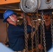 Two personnel in dark navy-style uniforms are working with a chain hoist in what appears to be an industrial or shipboard environment. The central figure wears a blue hard hat with chin strap and is operating or inspecting the chain hoist mechanism. A second individual with a mustache and dark cap observes from the right side.
