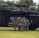 Four military personnel in camouflage uniforms are carrying a patient on a litter/stretcher away from a dark-colored military medevac helicopter marked with a red cross. The helicopter appears to be a UH-60 Black Hawk configured for medical evacuation, landed on a grass field. This appears to be either a training exercise or an actual medical evacuation operation.