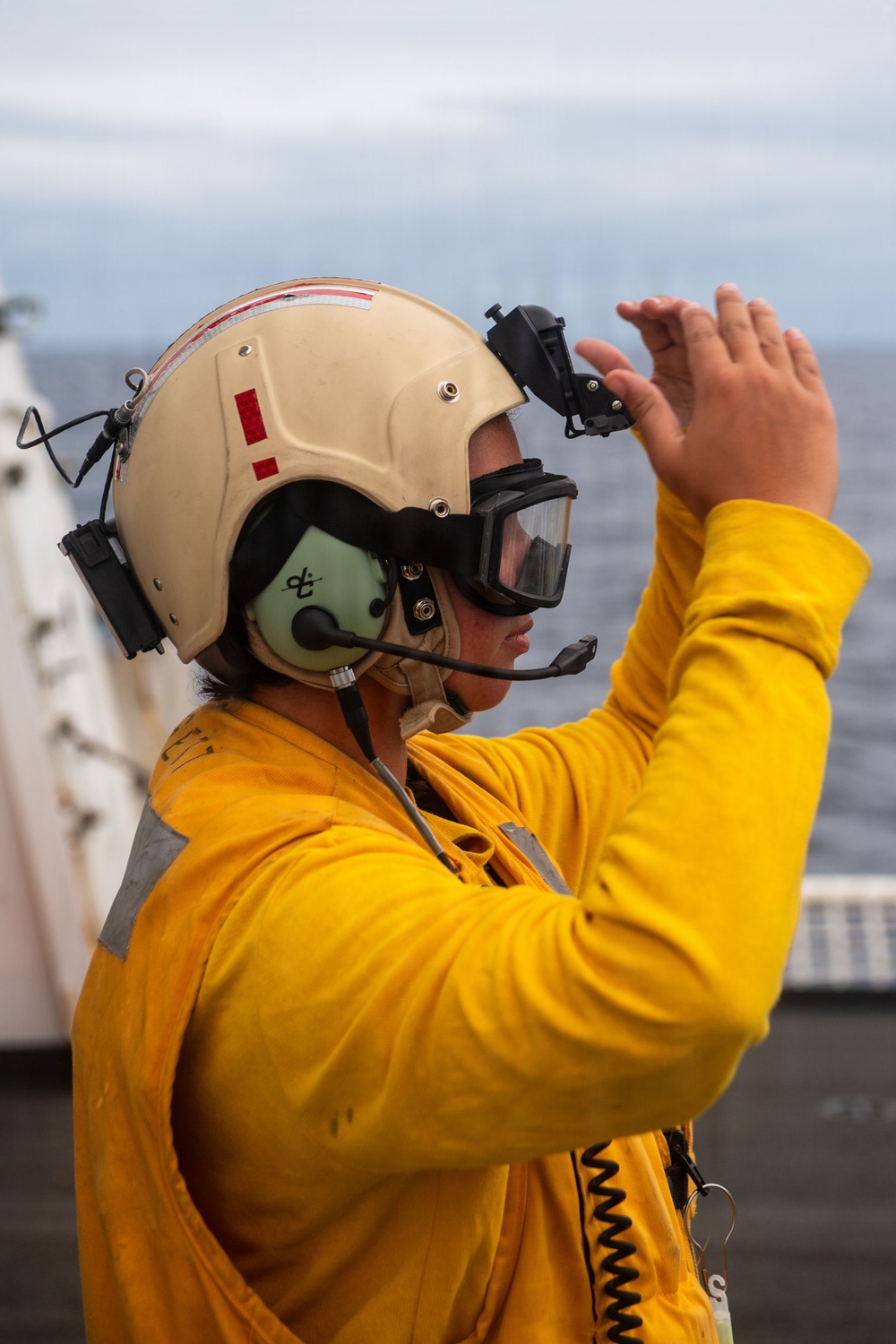 A naval flight deck crew member wearing a yellow jersey and tan helmet with communications equipment is performing hand signals on what appears to be an aircraft carrier flight deck. The yellow jersey indicates this individual is likely a aircraft handling officer or catapult/arresting gear officer. The ocean and overcast sky are visible in the background.