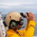 A naval flight deck crew member wearing a yellow jersey and tan helmet with communications equipment is performing hand signals on what appears to be an aircraft carrier flight deck. The yellow jersey indicates this individual is likely a aircraft handling officer or catapult/arresting gear officer. The ocean and overcast sky are visible in the background.