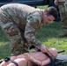 A U.S. Army soldier in OCP (Operational Camouflage Pattern) uniform is conducting a medical training exercise outdoors on a grass surface. The soldier is working with a CPR/medical training mannequin secured to a military litter (stretcher). A second soldier in matching uniform is visible in the background, suggesting a group training scenario.