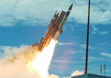 A missile is captured mid-launch from a ground-based launch facility, producing a large plume of white smoke and bright exhaust flame. The missile appears to have multiple booster stages attached and is angled diagonally upward against a partly cloudy sky. A small white utility building labeled '601A' is visible in the foreground right, with warning lights mounted on its roof.