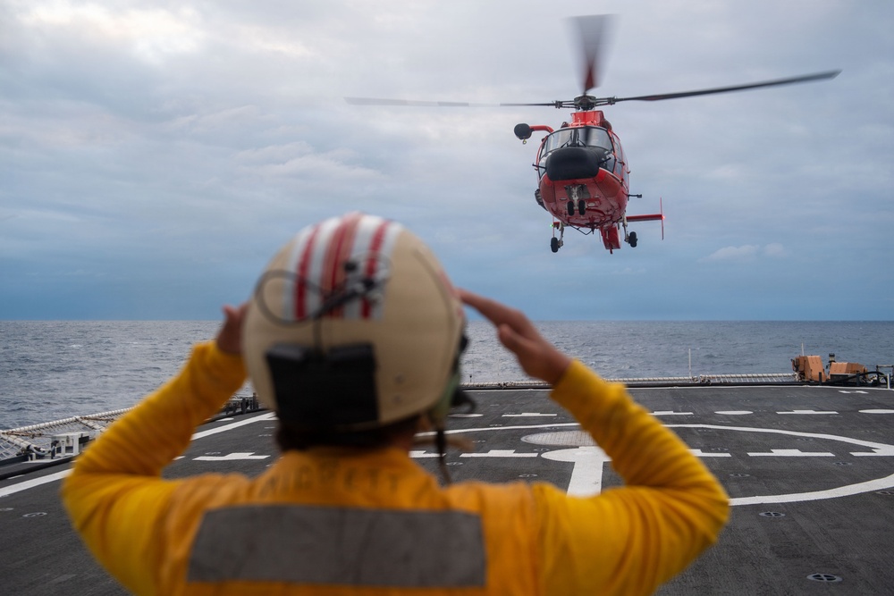 A flight deck crew member in a yellow jersey and helmet is guiding a red U.S. Coast Guard helicopter (MH-65 Dolphin) as it approaches or departs a ship's flight deck over open ocean. The crew member has both hands raised to their helmet, likely communicating via radio while signaling the aircraft. The scene depicts a standard helicopter deck operations procedure aboard a naval or Coast Guard vessel.