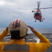 A flight deck crew member in a yellow jersey and helmet is guiding a red U.S. Coast Guard helicopter (MH-65 Dolphin) as it approaches or departs a ship's flight deck over open ocean. The crew member has both hands raised to their helmet, likely communicating via radio while signaling the aircraft. The scene depicts a standard helicopter deck operations procedure aboard a naval or Coast Guard vessel.