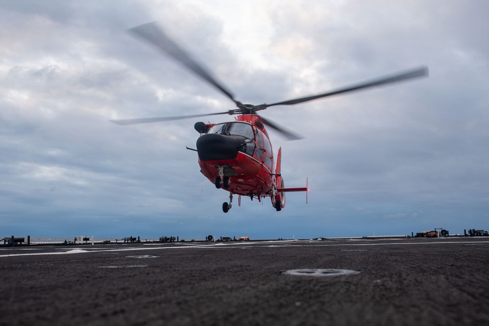 A red helicopter, consistent with a U.S. Coast Guard MH-65 Dolphin, is captured in a low hover or landing/takeoff phase above what appears to be a ship's flight deck. The rotor blades are visibly motion-blurred, indicating active rotation. The scene is set under an overcast, cloudy sky over open water.