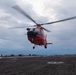 A red helicopter, consistent with a U.S. Coast Guard MH-65 Dolphin, is captured in a low hover or landing/takeoff phase above what appears to be a ship's flight deck. The rotor blades are visibly motion-blurred, indicating active rotation. The scene is set under an overcast, cloudy sky over open water.