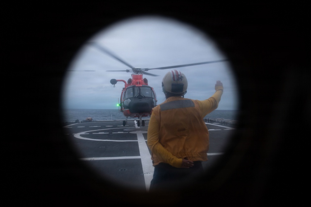 The image is captured through a circular porthole or viewport, showing a ship's flight deck with a red helicopter (consistent with U.S. Coast Guard livery) landed or hovering on the helipad. A deck crew member wearing a yellow jersey and flight deck helmet is visible from behind, raising one arm in a signaling gesture toward the aircraft. The scene takes place at sea under overcast skies.