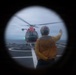 The image is captured through a circular porthole or viewport, showing a ship's flight deck with a red helicopter (consistent with U.S. Coast Guard livery) landed or hovering on the helipad. A deck crew member wearing a yellow jersey and flight deck helmet is visible from behind, raising one arm in a signaling gesture toward the aircraft. The scene takes place at sea under overcast skies.
