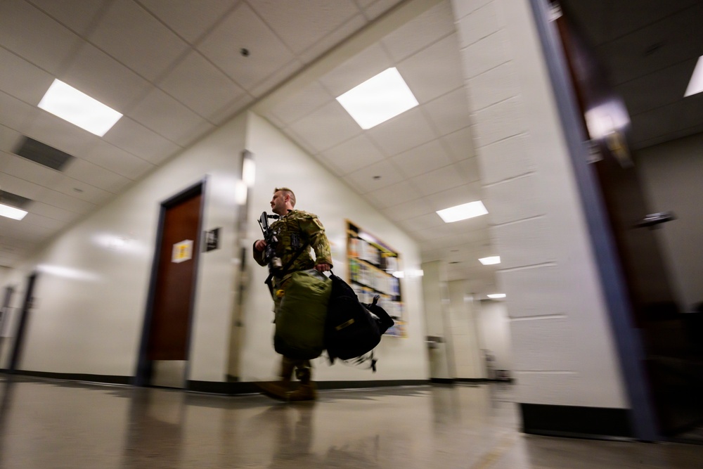 A soldier in military camouflage uniform is moving rapidly through an indoor corridor, carrying large duffel bags. The photo is taken from a low angle with motion blur, suggesting urgency or rapid movement. The setting appears to be a institutional building such as a school or government facility based on the hallway design.