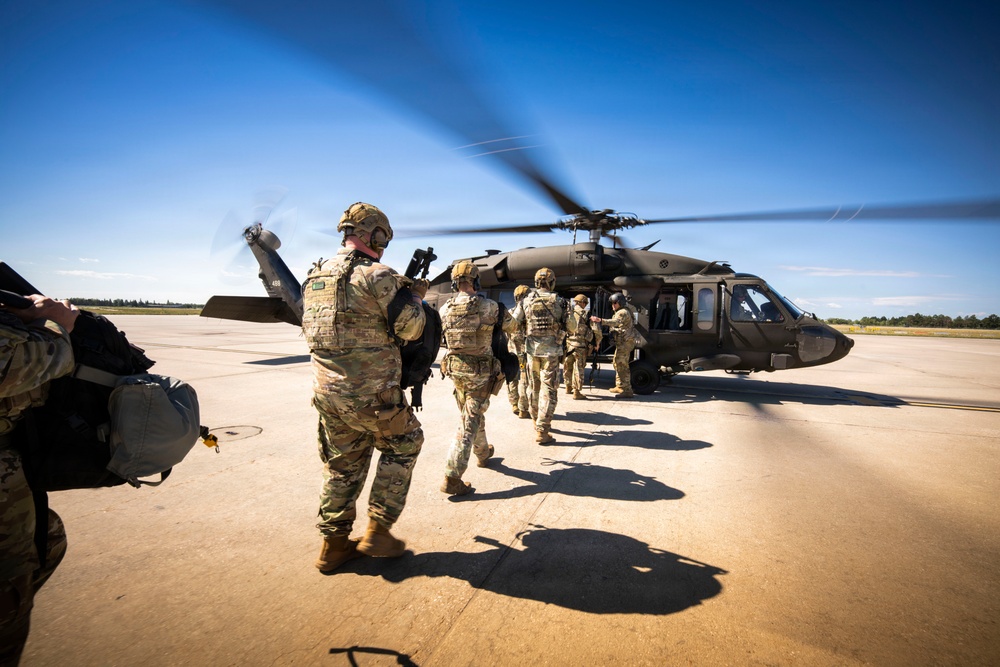 A group of military personnel in full combat gear are boarding or approaching a UH-60 Black Hawk helicopter on a tarmac or airfield. The helicopter's rotors are spinning, indicating it is active and ready for departure. The scene appears to depict a military air assault or troop insertion exercise.