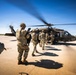 A group of military personnel in full combat gear are boarding or approaching a UH-60 Black Hawk helicopter on a tarmac or airfield. The helicopter's rotors are spinning, indicating it is active and ready for departure. The scene appears to depict a military air assault or troop insertion exercise.