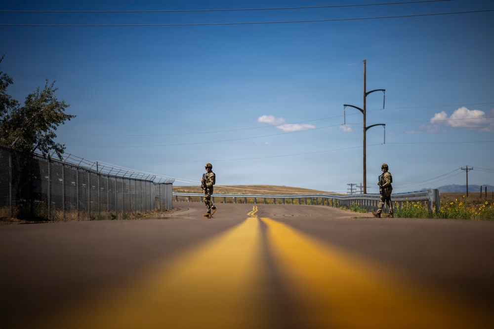 Two armed soldiers in camouflage uniforms and helmets stand on a two-lane road with yellow center lines, appearing to form a checkpoint or patrol position. A tall security fence with razor wire runs along the left side of the road, suggesting a secured perimeter or border area. The photo is taken from a low ground-level angle, emphasizing the road's perspective lines leading toward the soldiers.