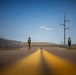 Two armed soldiers in camouflage uniforms and helmets stand on a two-lane road with yellow center lines, appearing to form a checkpoint or patrol position. A tall security fence with razor wire runs along the left side of the road, suggesting a secured perimeter or border area. The photo is taken from a low ground-level angle, emphasizing the road's perspective lines leading toward the soldiers.